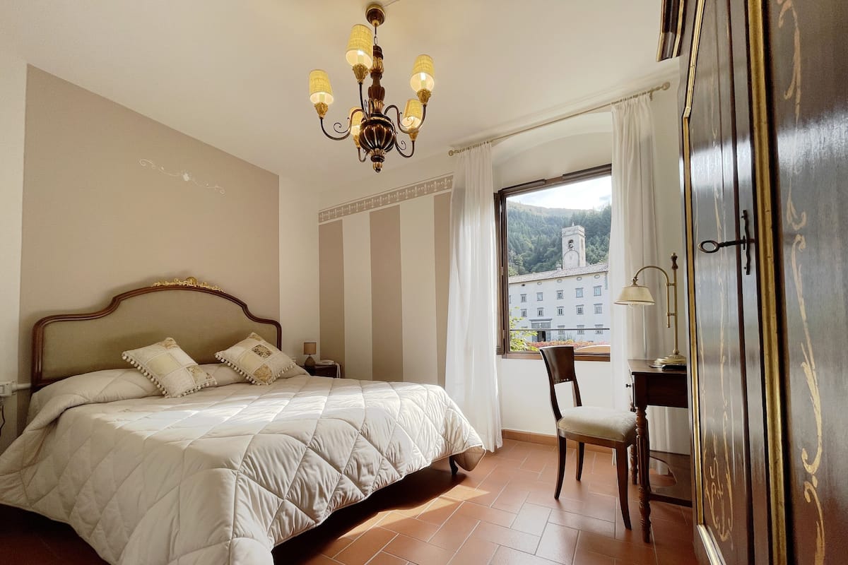 Une chambre d’hôtel avec un grand lit, un bureau, une chaise, un lustre et une vue sur un immeuble par la fenêtre.