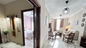 Ein geräumiges Wohnzimmer mit einem großen weißen Sofa, zwei Schaukelstühlen, einem Couchtisch und einem Fernseher. Ein Durchgang führt in ein Schlafzimmer mit Bett und Deckenventilator. Der Raum hat einen gefliesten Boden und einen Kronleuchter.
