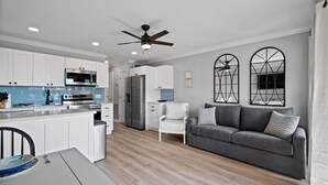 Una cocina moderna con armarios blancos, un microondas y un horno empotrado. En la sala de estar, hay un sofá gris y una silla blanca.