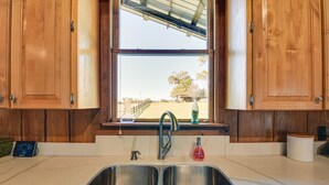 Una cocina con armarios de madera, un lavamanos doble, una ventana con vista a un jardín y un reloj digital.