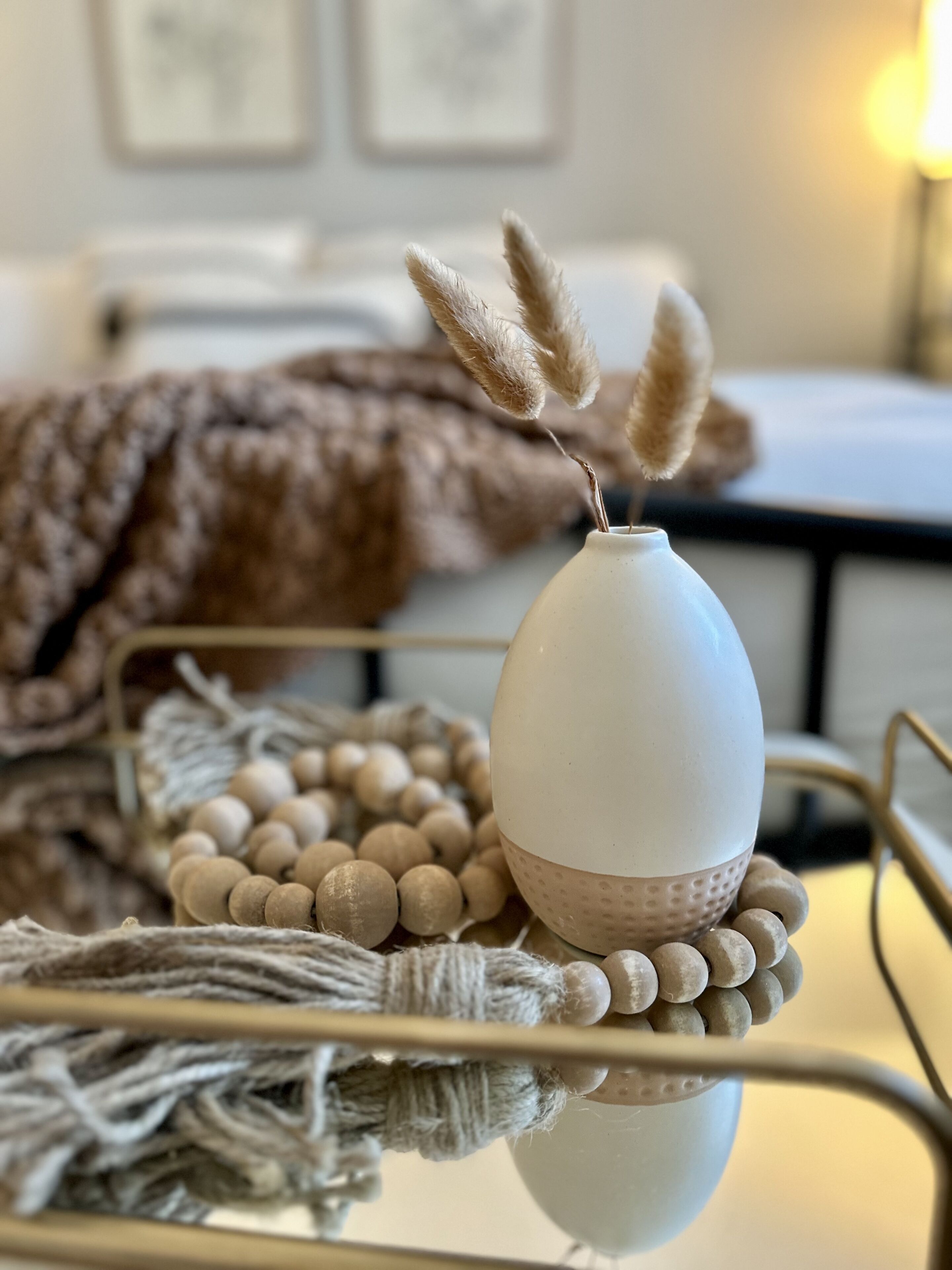 Un vaso bianco con pampas secchi su un vassoio metallico con base intrecciata, circondato da perline di legno e un elemento in macramè.