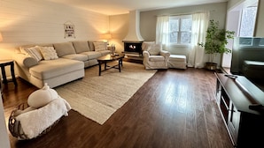 A living room with a sectional sofa, a fireplace, a TV, and a large window with curtains.