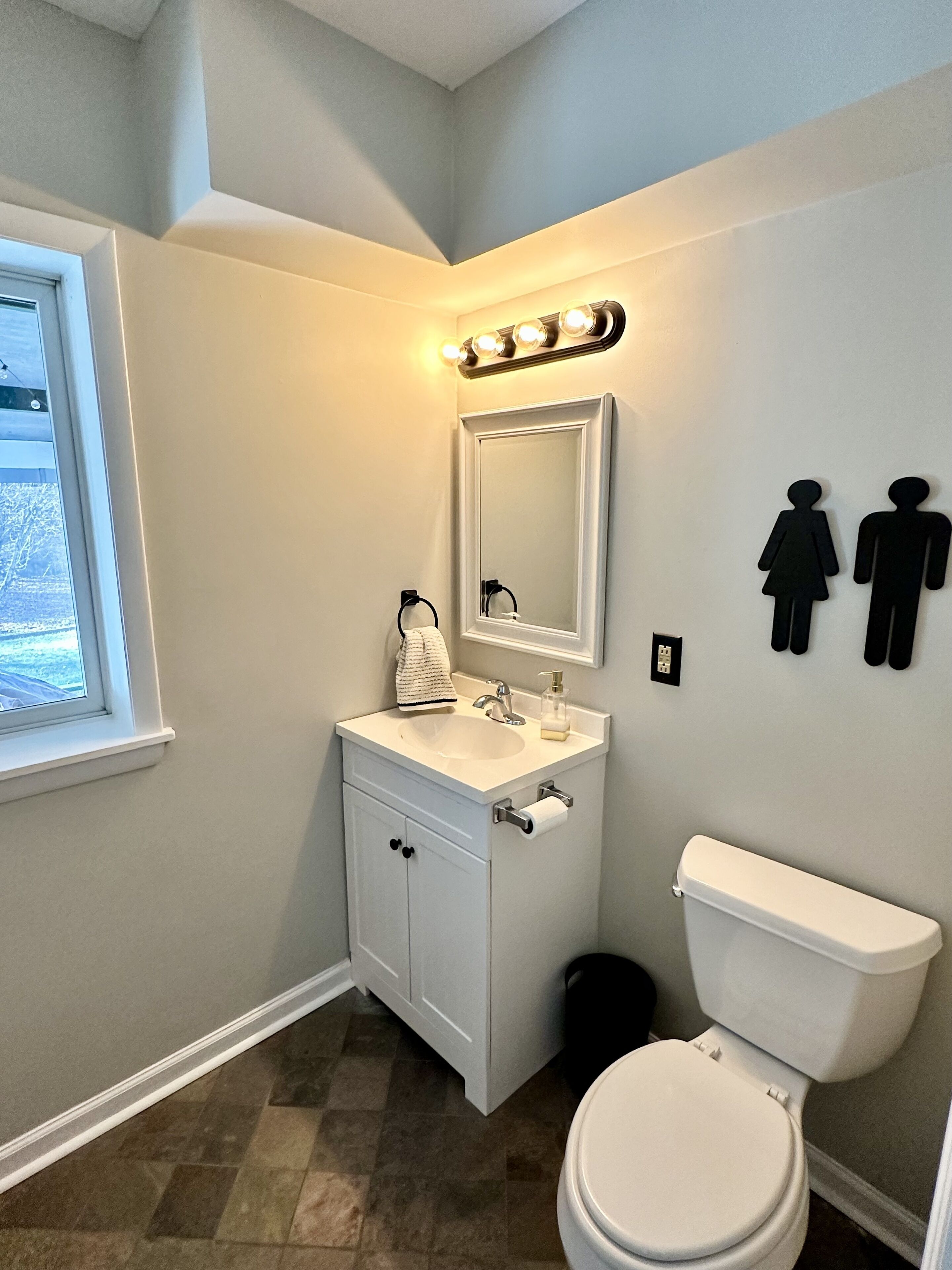 Un bany amb un lavabo blanc, un mirall i un vàter. Hi ha llums penjades a la paret sobre el mirall i dos símbols de gènere a la paret.