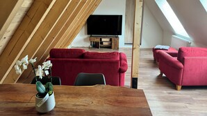 A modern loft with a sloped ceiling, wooden beams, a red sofa, a wooden dining table, and a potted orchid.