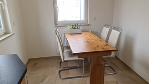Quarto pequeno e limpo, com uma mesa de madeira e cadeiras brancas, uma planta em vaso e uma janela com vista.