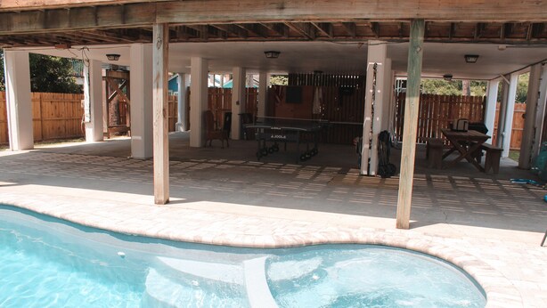 A covered outdoor swimming pool with a hot tub, wooden deck, and barbecue area.