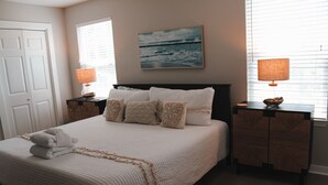 A neatly made bed with a white comforter and decorative pillows, a painting of a seascape on the wall, and two bedside tables with lamps.