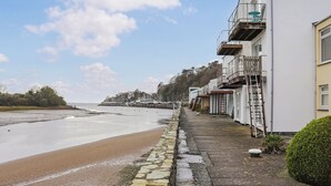 Un bâtiment situé sur la côte, avec un sentier de pierre menant à la plage, une échelle sur le côté et une vue sur la mer et les arbres au loin.