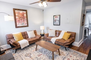 A living room with two brown leather sofas, a wooden coffee table, a patterned rug, a ceiling fan, and wall art.