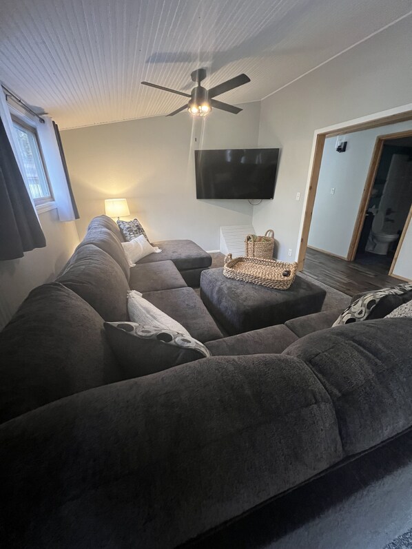 A living room with a grey sectional sofa, a ceiling fan, a flat-screen TV, and a basket on a ottoman.