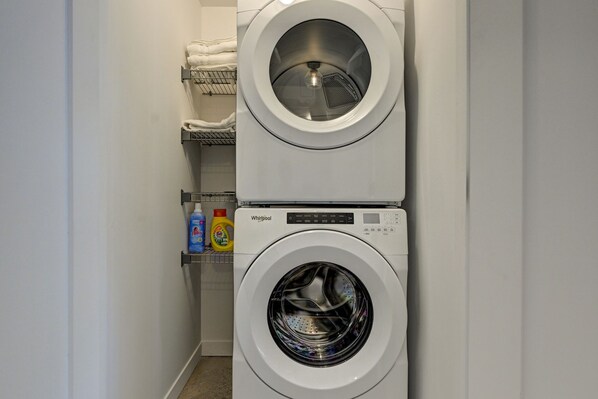 Une machine à laver et un sèche-linge superposés dans une buanderie, avec des étagères au-dessus où sont rangées des serviettes pliées et du matériel de lessive.