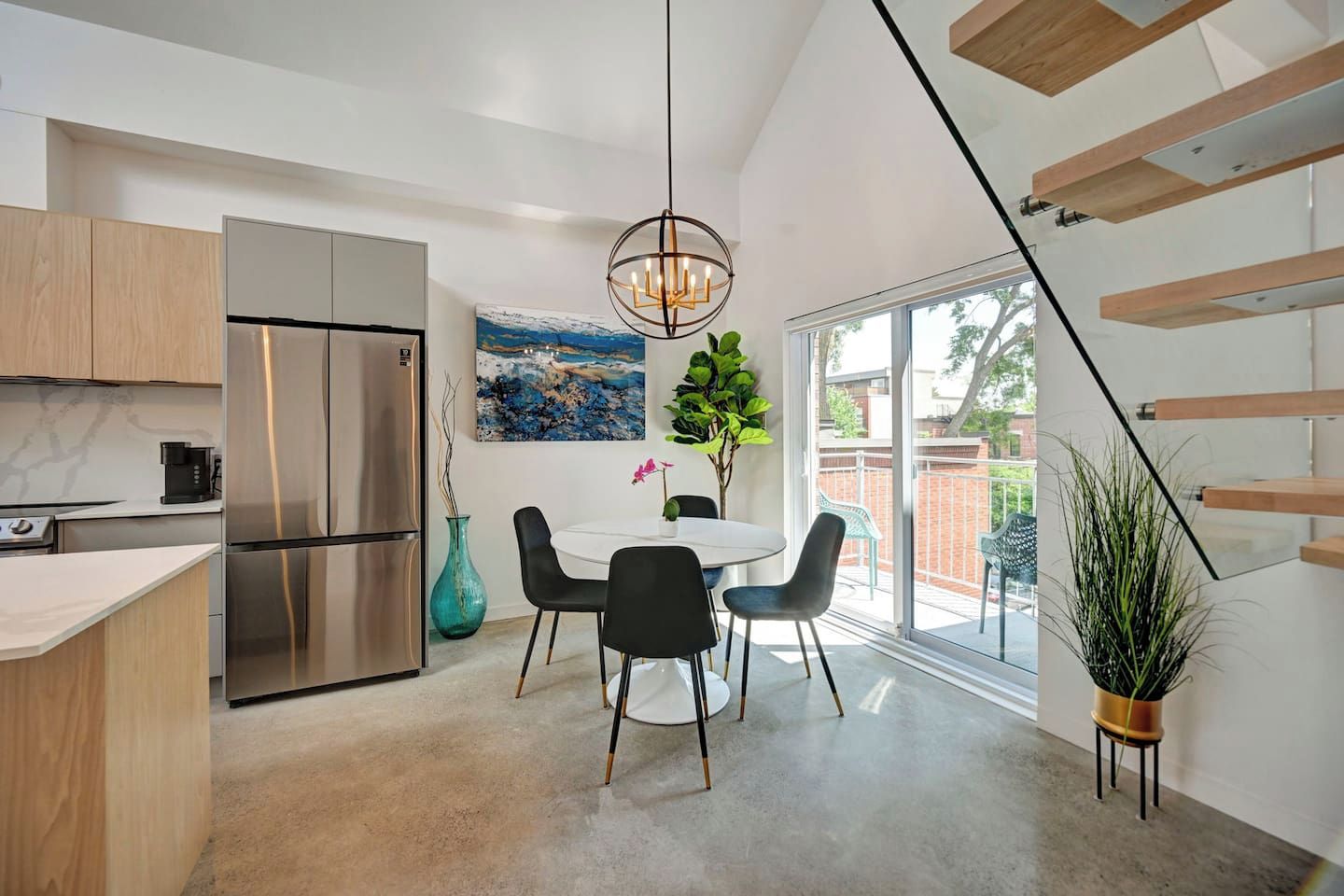 Una cocina moderna con un refrigerador de acero inoxidable, una zona de comedor con una mesa blanca y sillas negras, y un ventanal con vistas al exterior.
