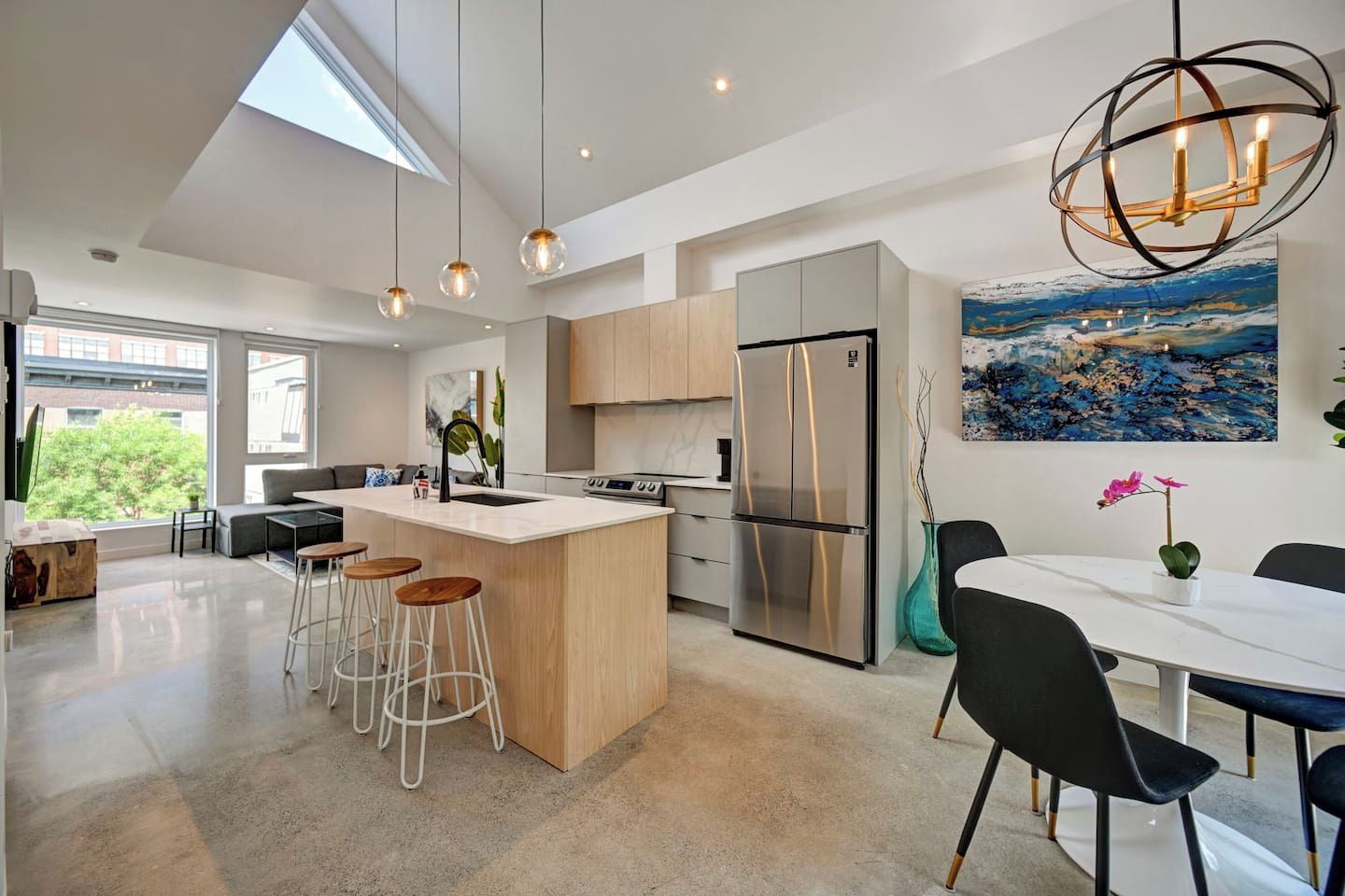 Una cocina moderna con una isla central, taburetes de bar, una mesa de comedor con sillas, un refrigerador y un cuadro en la pared.