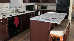 Une cuisine moderne avec des meubles de couleur bois foncé, un îlot central doté de tabourets, un réfrigérateur noir et une fenêtre avec des stores.