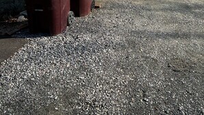Une maison avec une allée en gravier menant à un porche blanc, deux poubelles et une vue sur un lac.