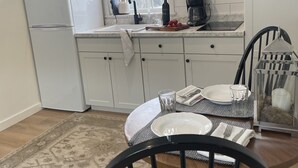 Une cuisine compacte avec des meubles blancs, un réfrigérateur, un four à micro-ondes et une machine à café. On y trouve une table à manger avec des chaises, un tapis et une lanterne décorative.