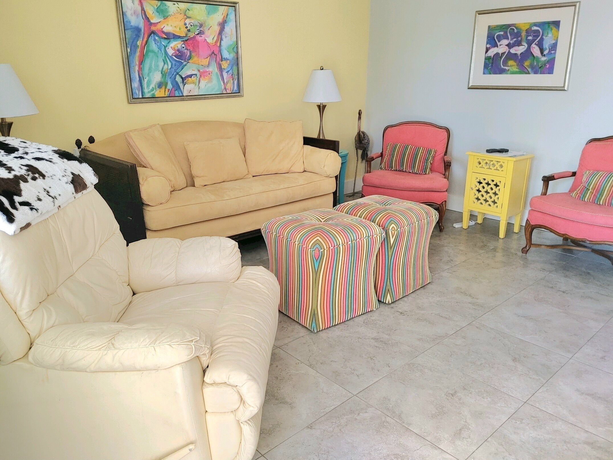 A living room with a beige sofa, a pink armchair, and a striped ottoman. There are two lamps, a small table, and a painting on the wall.
