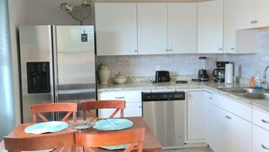 A modern kitchen with white cabinets, a stainless steel refrigerator, a dishwasher, and a dining area with wooden chairs and a table.
