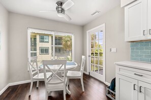A kitchen with white cabinets, a white dining table with chairs, a ceiling fan, and a view of the outdoors through the windows.