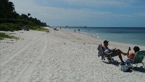 A wide sandy beach with people relaxing on chairs, a clear sky, and a distant pier.
