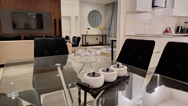 A modern living room with a glass coffee table, black chairs, a flat-screen TV, and a dining area with a white table and chairs. - Victor Regents Apartment (Accra)
