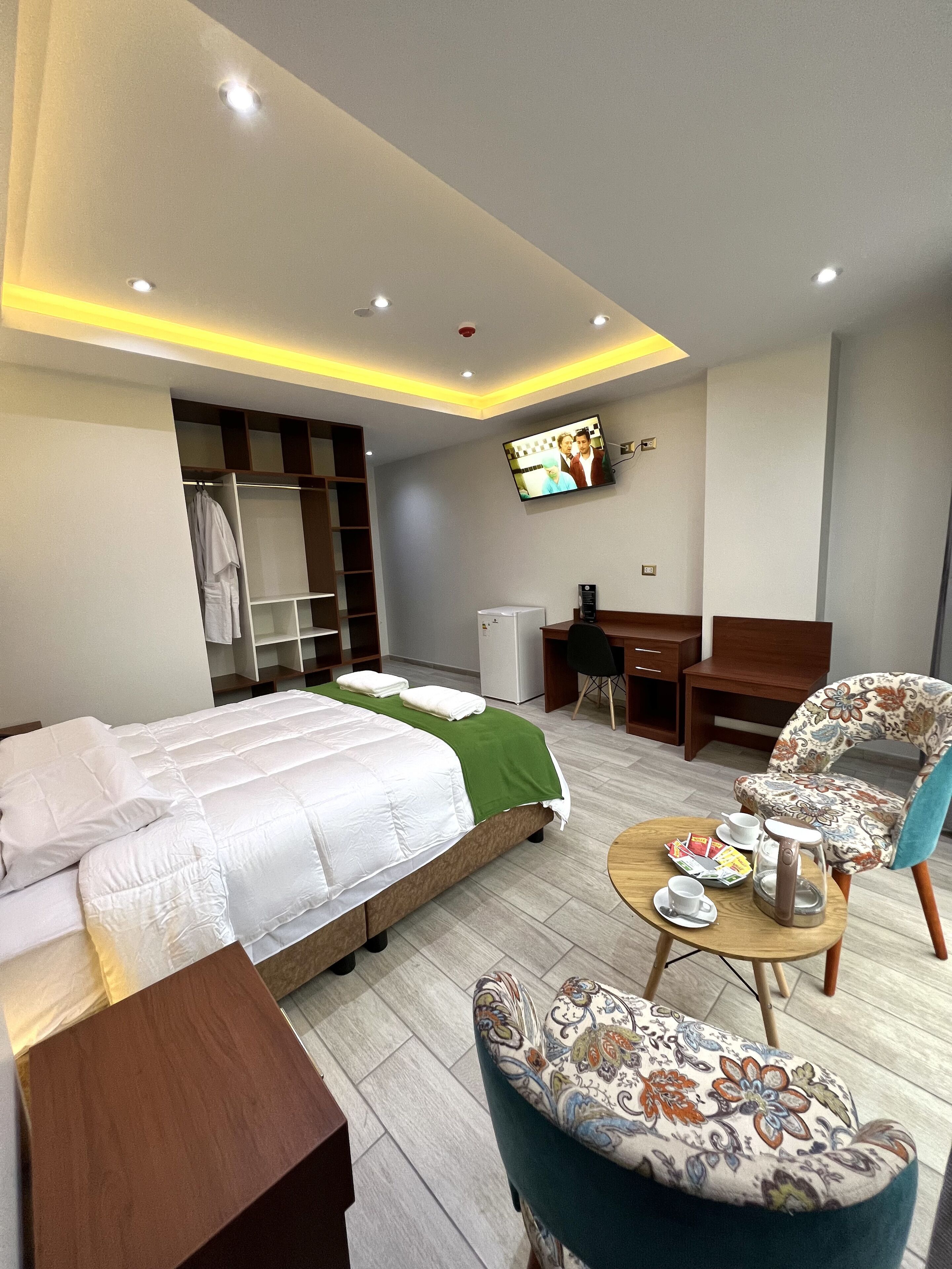 A hotel room with a bed, a desk, a chair, a television, and a small table with a kettle and cups.