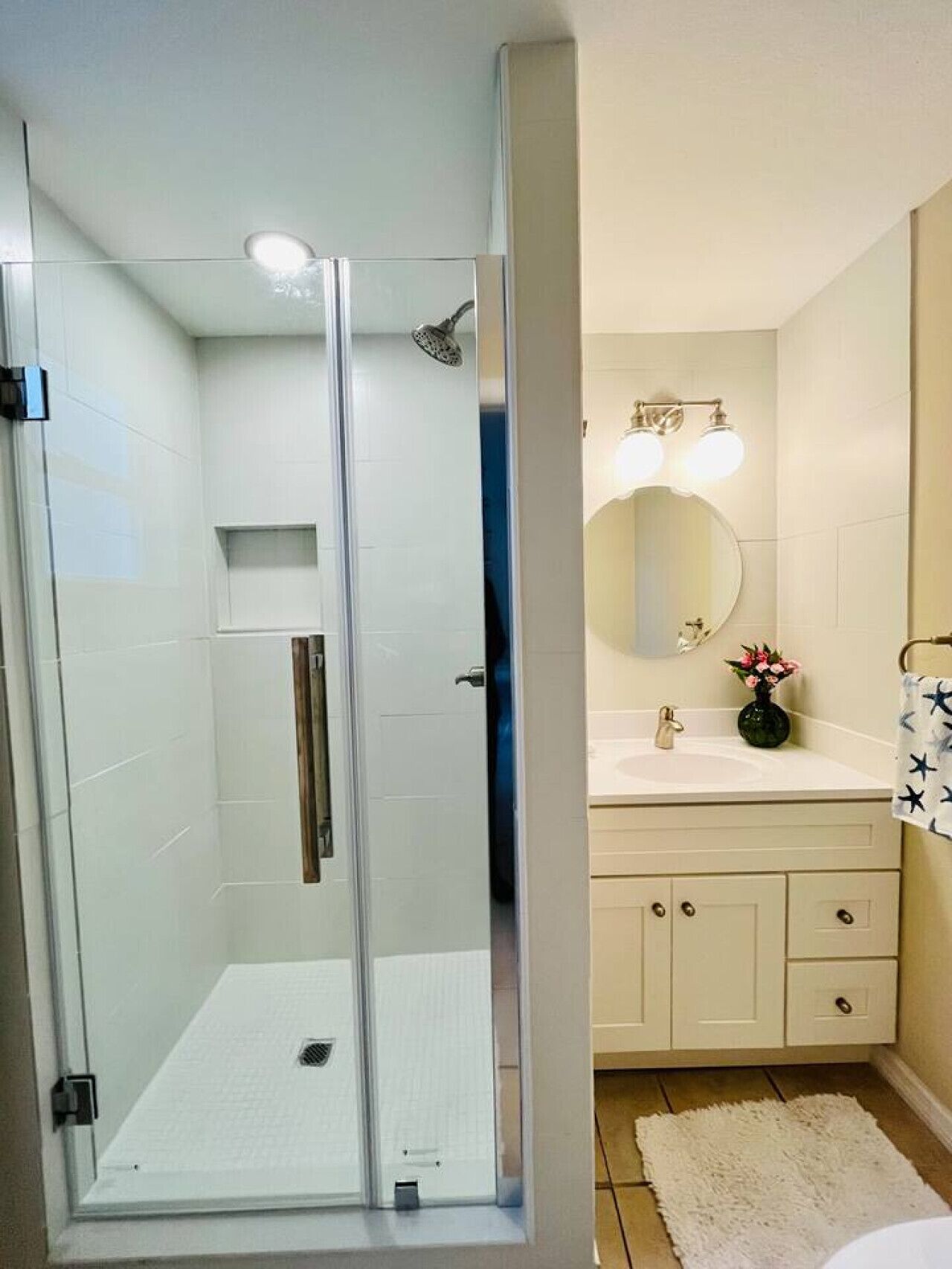 A bathroom with a glass-enclosed shower, a sink with a mirror, and a small vase with flowers.