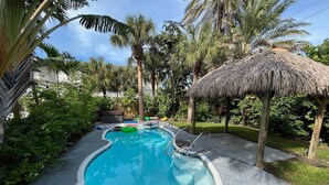 A swimming pool with a thatched roof structure, surrounded by palm trees and greenery. - `Dunes` Gulf-View Historic Beachside Cottage 1/1 (Indian Rocks Beach)