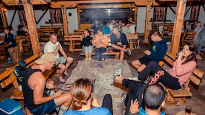 A group of people seated around a stone fire pit in a rustic wooden room with benches and tables. - Camp Divlja Rijeka (Foca)