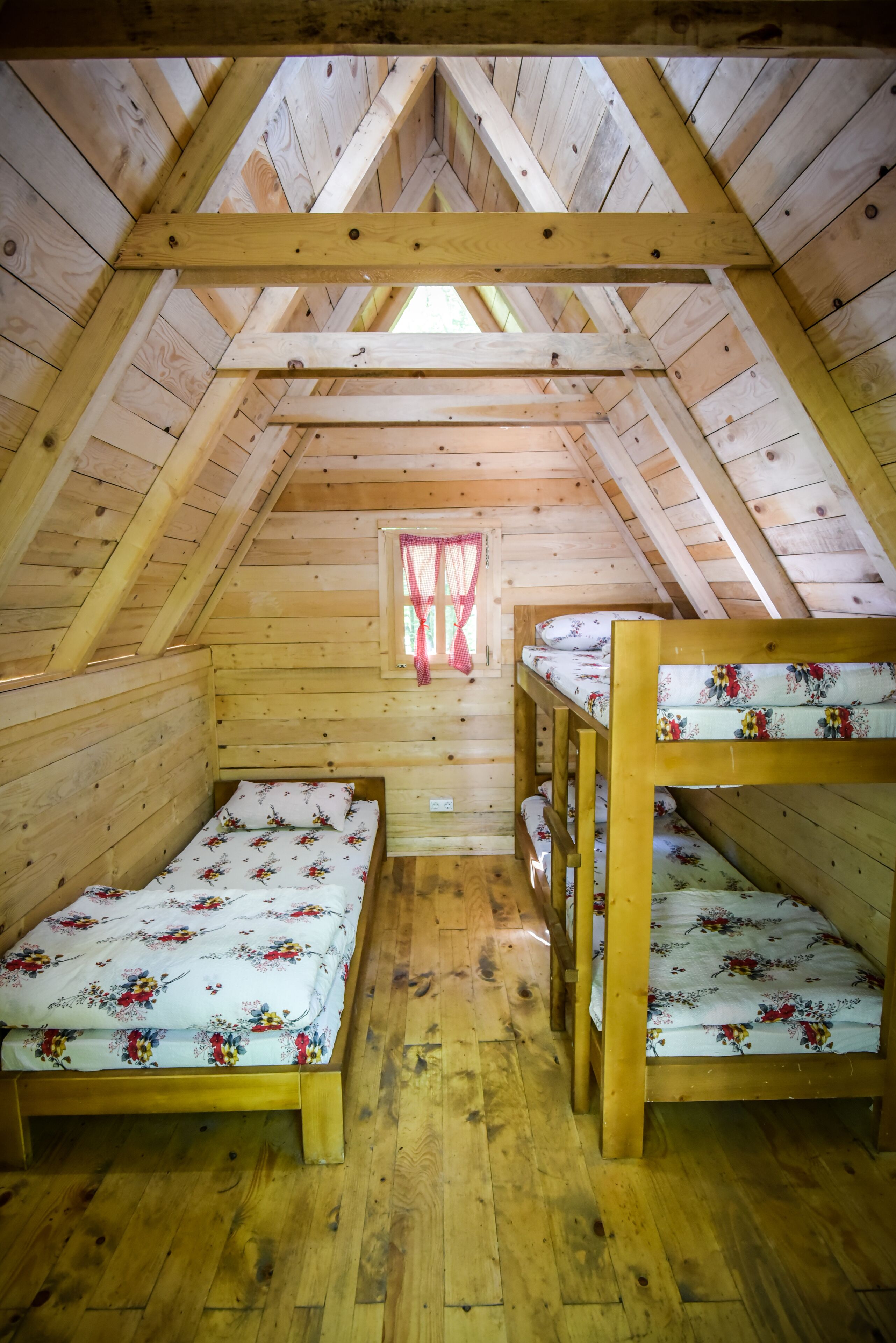 A small attic bedroom with wooden walls and a sloped ceiling. There are two bunk beds with floral bedding and a window with red curtains.
