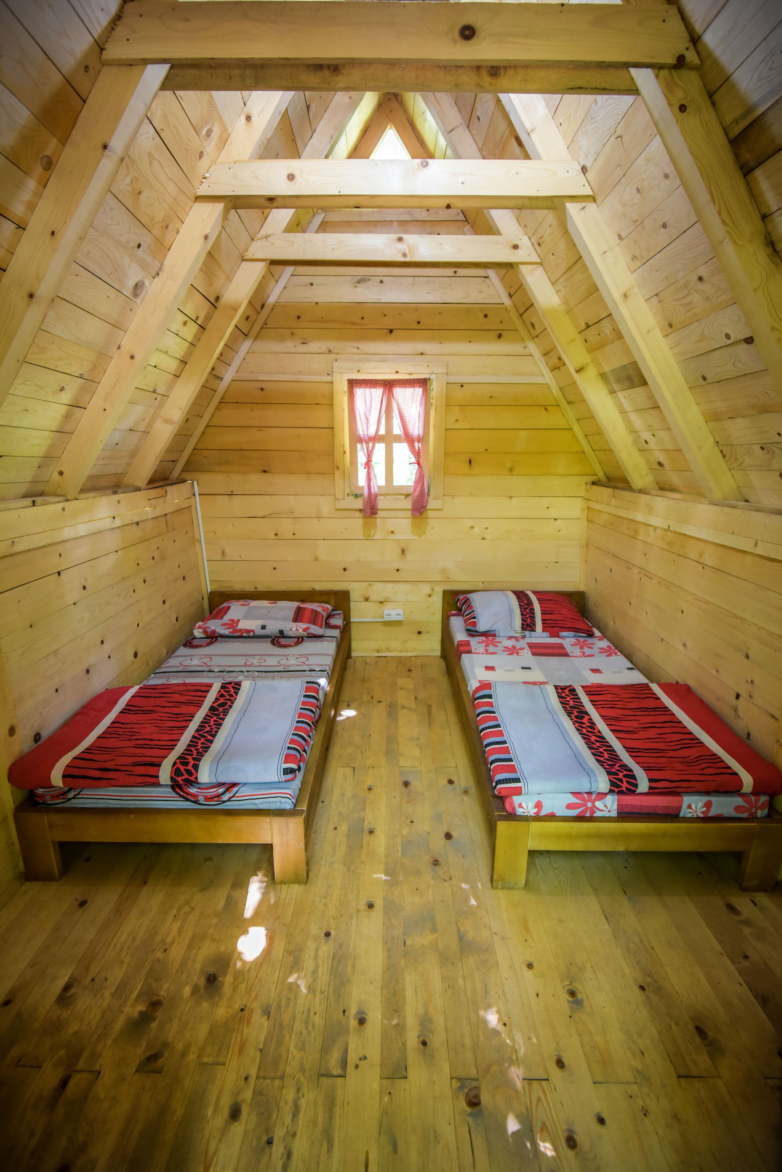 A small, wooden attic bedroom with two single beds, a window with curtains, and a sloped ceiling.