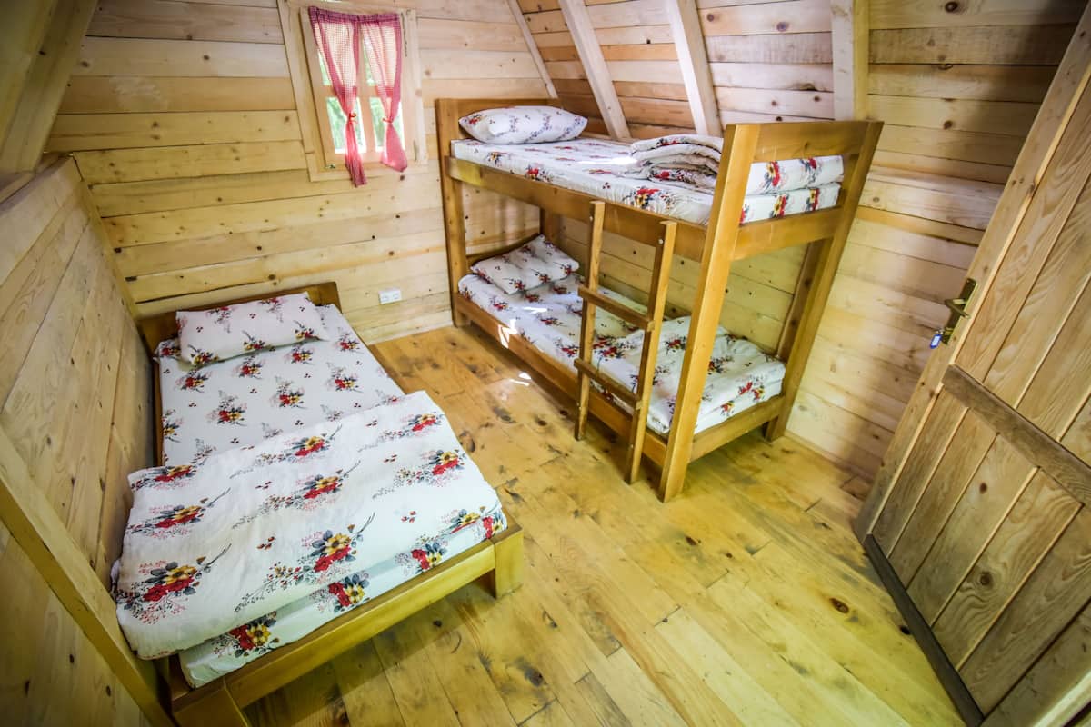 A wooden cabin room with bunk beds and a single bed, wooden floors, and a window with a red curtain.