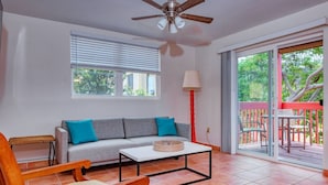 A living room with a sofa, a coffee table, a chair, a ceiling fan, a lamp, and a sliding glass door leading to an outdoor deck.