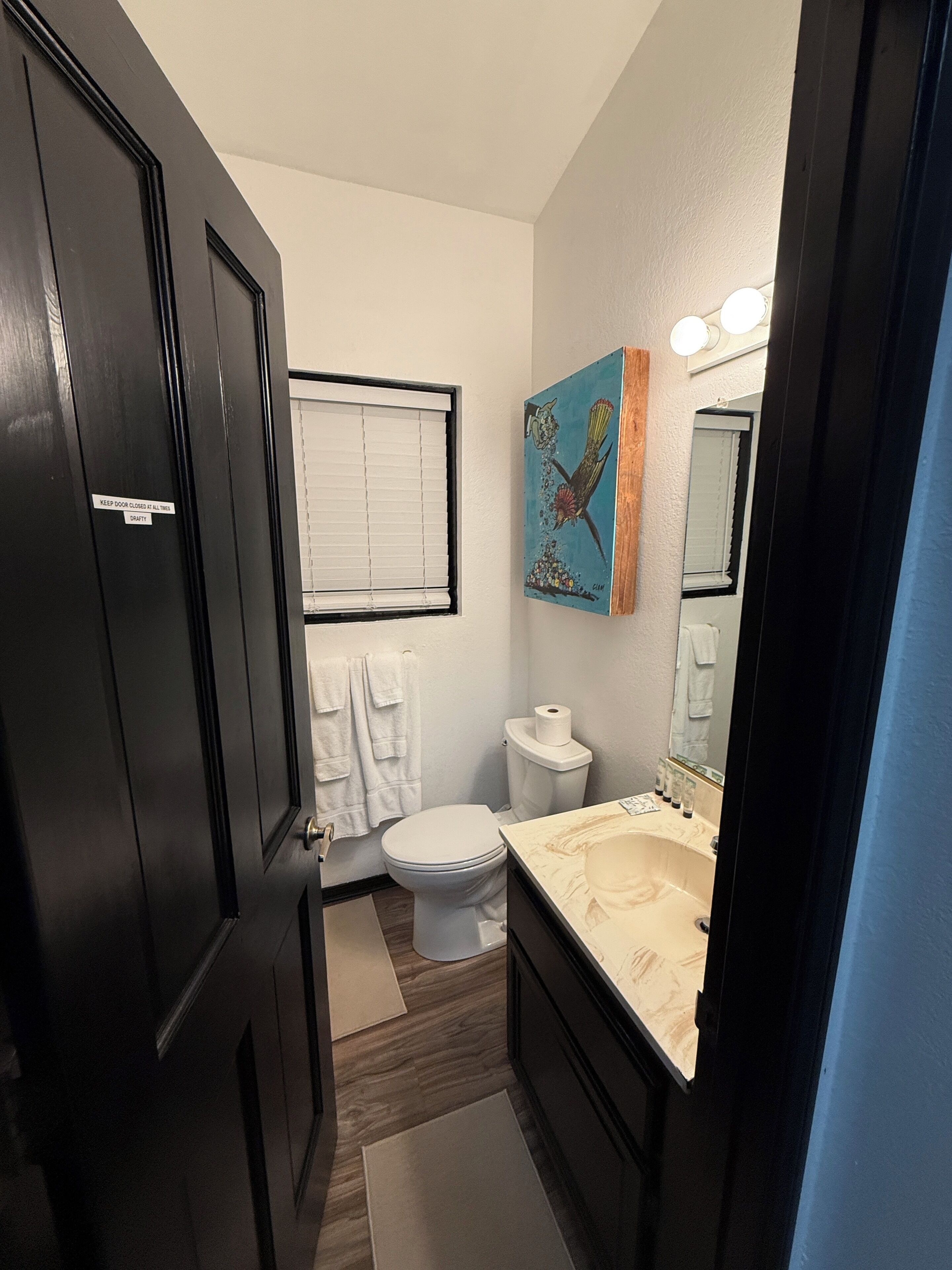Ein Badezimmer mit Toilette, Waschbecken und Spiegel. Handtücherhalter mit Handtüchern, ein Bild an der Wand und eine Tür mit einem Schild.