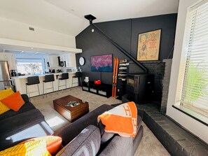 Una sala de estar moderna con chimenea, una cocina al fondo y una escalera que sube a un nivel superior.