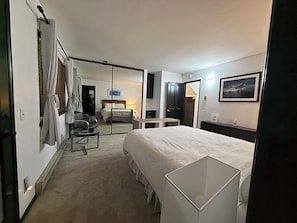 Ein Hotelzimmer mit Bett, Schreibtisch, Stuhl, Kleiderschrank und einem durch eine Glaswand erreichbaren Badezimmer.