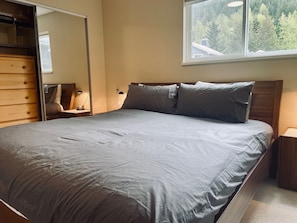 A bedroom with a bed, wooden dresser, and a window with a view of trees.