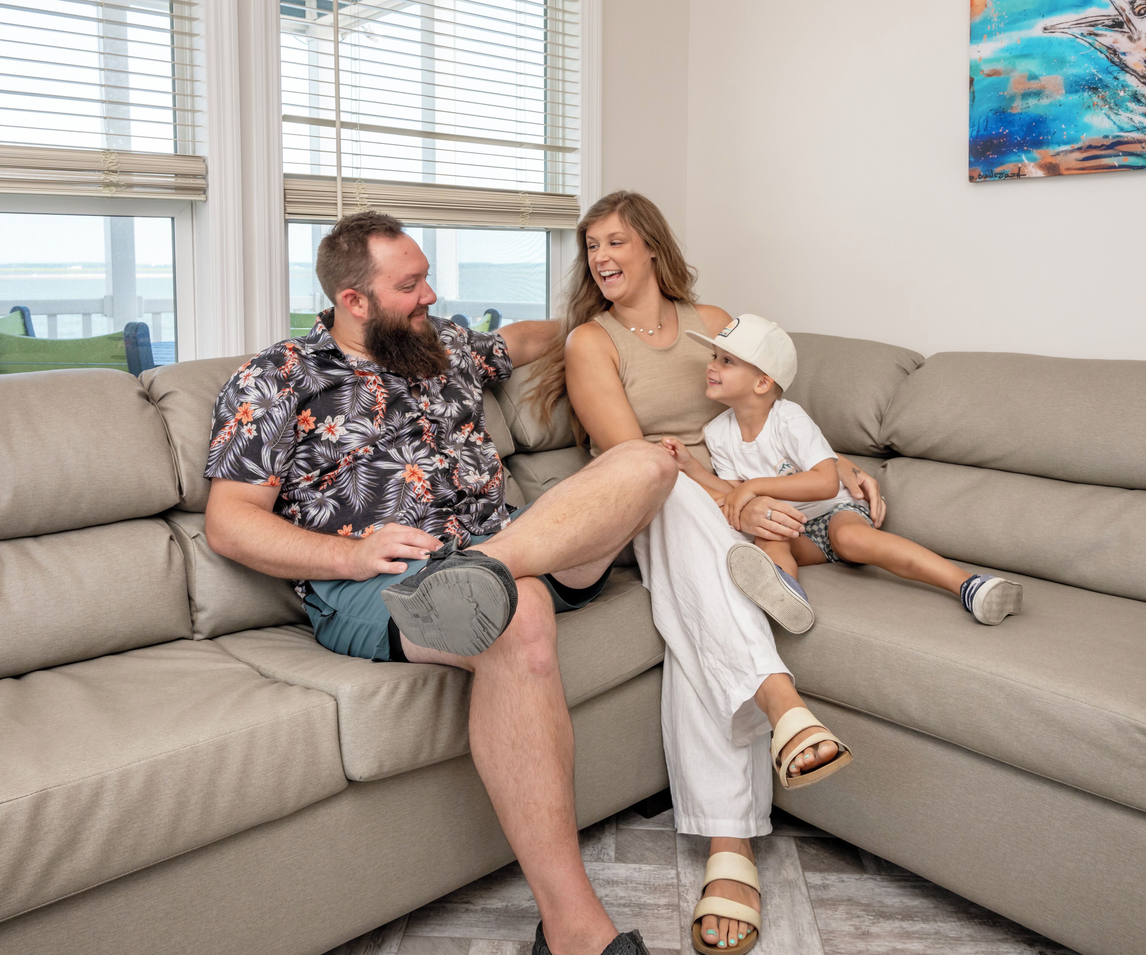 A family sitting on a beige sectional sofa in a living room with a large window and a painting on the wall.