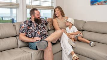 A family sitting on a beige sectional sofa in a living room with a large window and a painting on the wall.