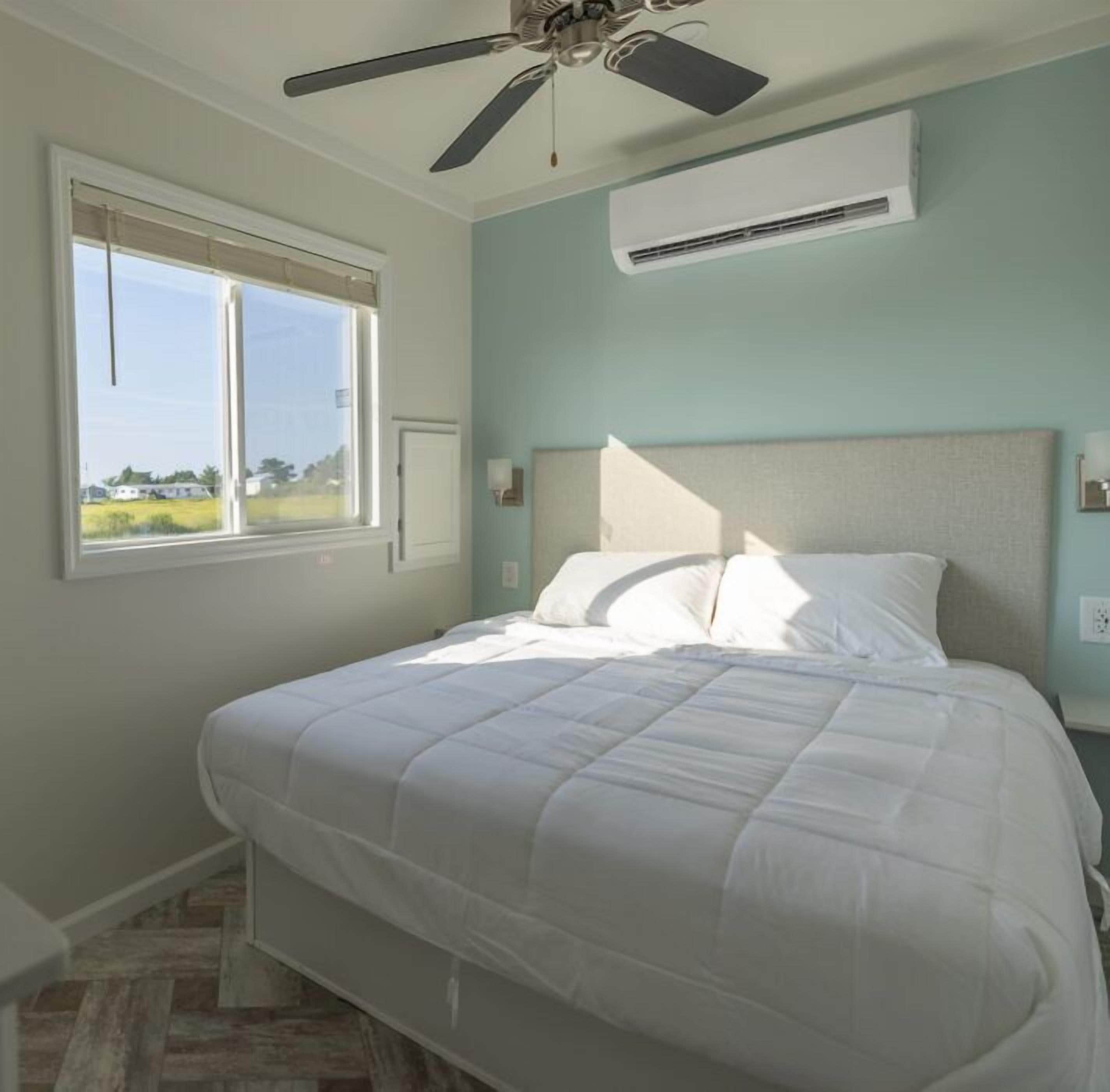 A bedroom with a bed, a ceiling fan, an air conditioning unit, a window, and a view of the outdoors.