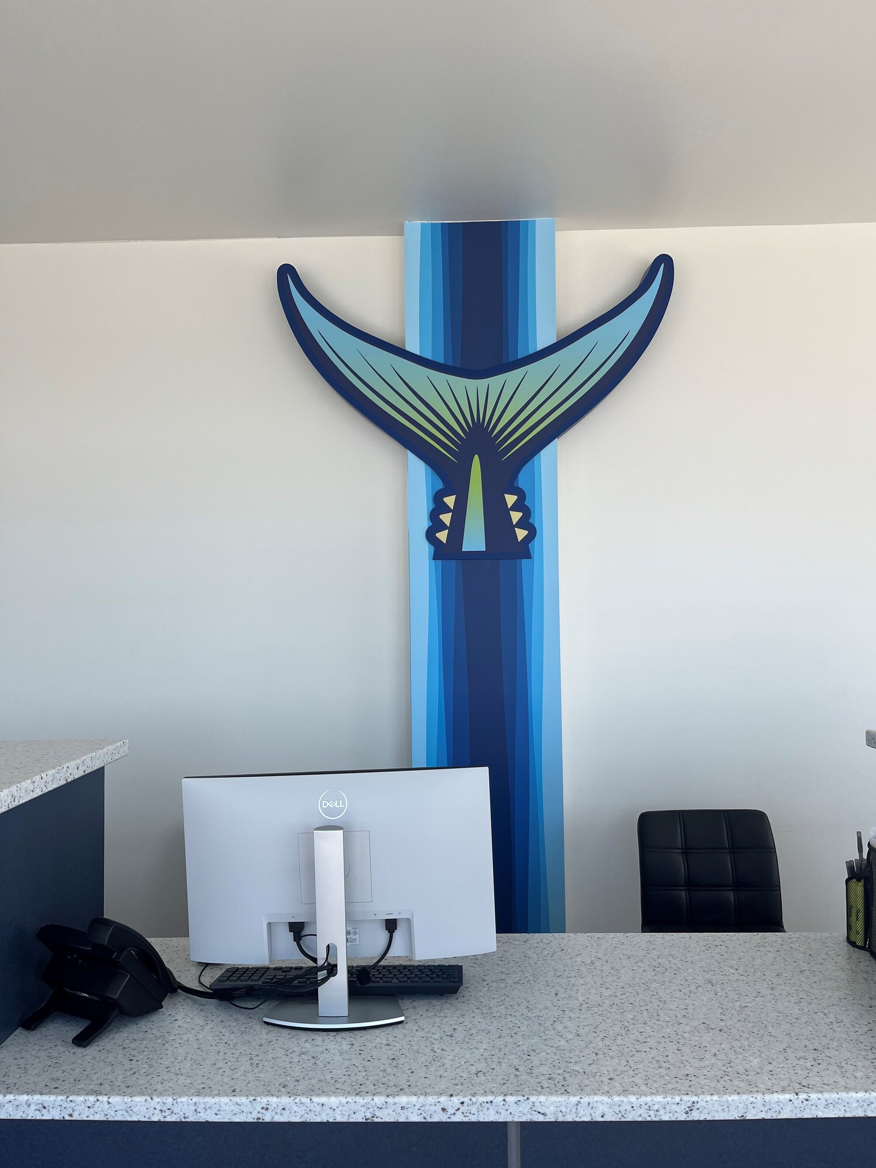 Bancada de recepção com monitor Dell, telefone e teclado. Há uma cadeira de escritório preta e parede com um desenho abstrato em azul e verde.