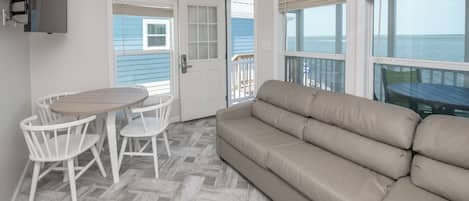 A room with a beige sofa, a wooden table with white chairs, and a view of the ocean through large windows.