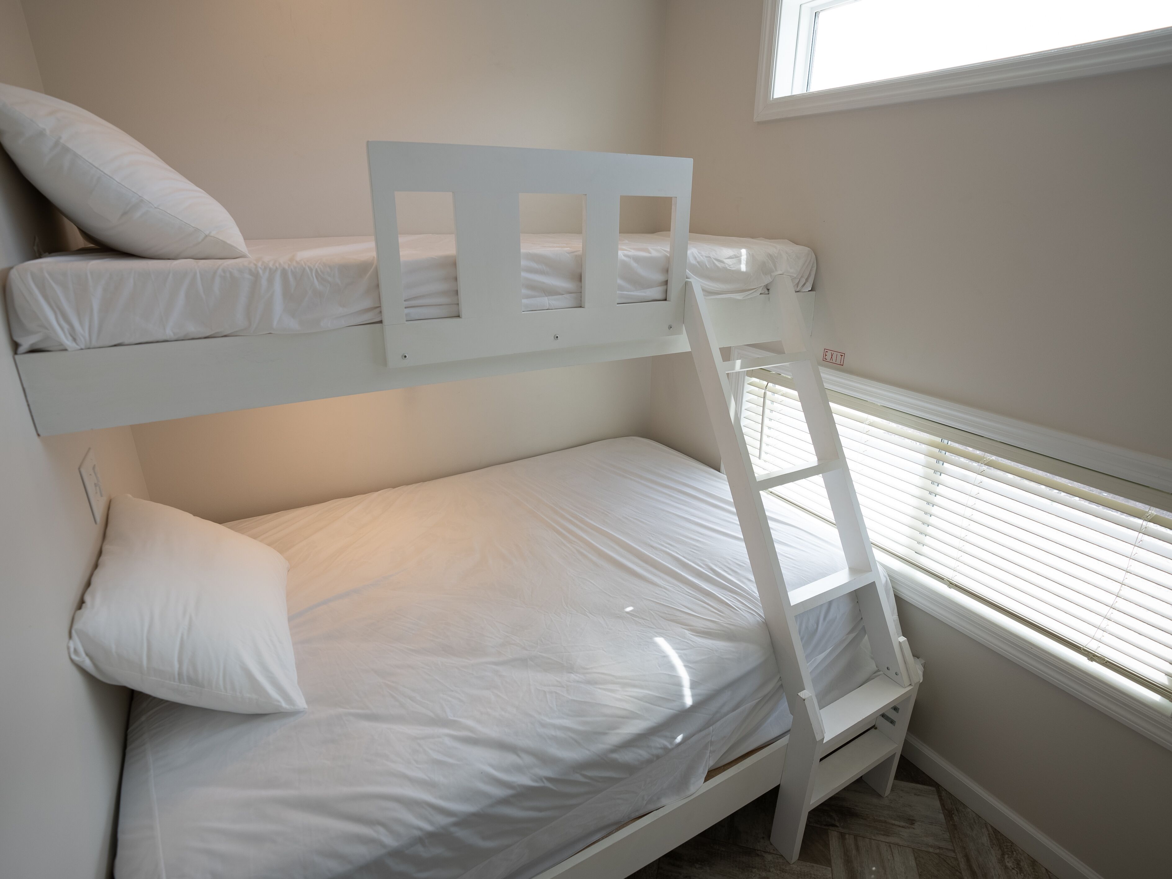 A bunk bed with white bedding and a ladder, a window with blinds, and a wall-mounted light switch.