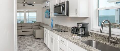 A modern kitchen with white cabinets, a granite countertop, a built-in microwave, a coffee maker, and a large window overlooking the sea.