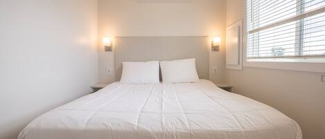A neatly made bed with white bedding, a ceiling fan, an air conditioning unit, and two wall-mounted lamps.