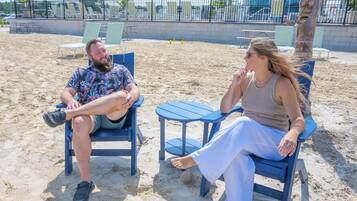 Two people seated on beach chairs with a small table between them, on a sandy beach with umbrellas and a building in the background.