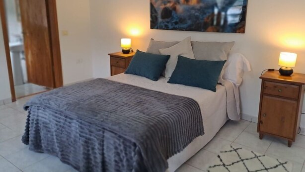 A neatly made bed with a gray blanket, two bedside tables with lamps, and a painting of a coastal scene above the bed. - Lovely Private House in Taganana - Anaga Rural Park (Santa Cruz de Tenerife)