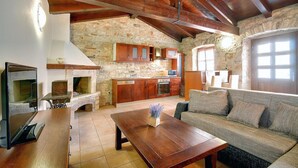 A rustic interior with exposed stone walls, wooden beams, a fireplace, a kitchen with wooden cabinets, and a seating area with a sofa and coffee table.