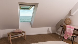 Une chambre avec un plafond en pente, une fenêtre donnant sur la mer, une chaise en bois, une petite table sur laquelle repose un panier en osier, et un lit avec une serviette rose.