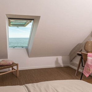Una habitación con techo inclinado, un ventanal con vista al mar, una silla de madera y una cama.